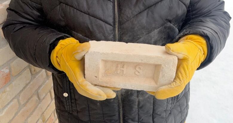 A person wearing yellow gloves holds a brick with the initials HS.