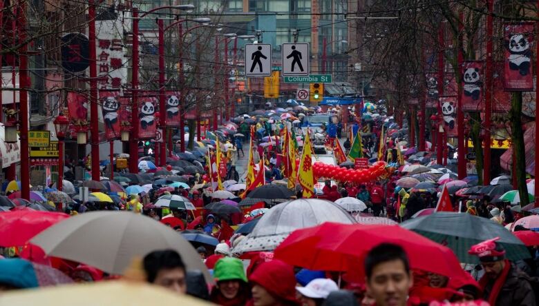 A substantial crowd of people hold umbrellas standing on the street, with trees and panda-featuring banners on both sides of the street and dragon dancers in the background.