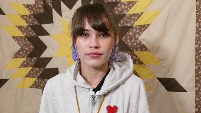 A young woman with brown hair in a ponytail and an off white sweatshirt standind in front of a tapestry quilt.