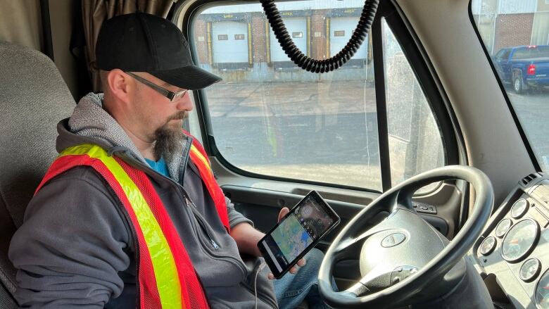 A man in a high visibility vest is sitting in the driver's seat of a truck looking at an electronic logging device showing his route. 