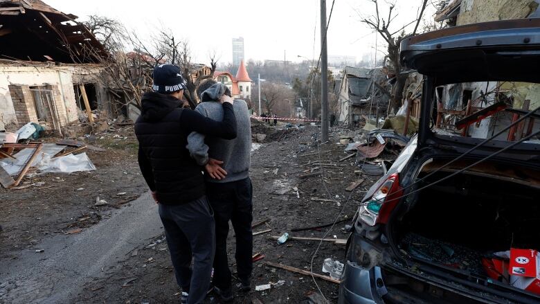 Two people hug outside next to homes and vehicles that have been badly damaged. 