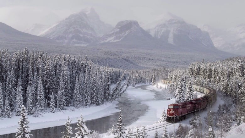 A red train travels on a winding railway through snowy trees near an icy Bow River while the mountains appear in a snowy fog. 