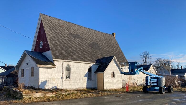 A small white church building with a sloped roof is next to a parking lot that contains a power lift to repair the roof.