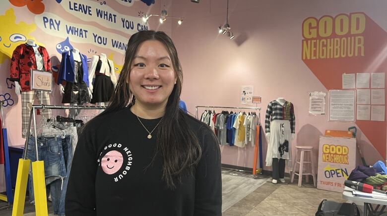 A woman stands in a retail store smiling for the camera. She is wearing a black sweater with a logo reading 'Good Neighbour'. The store's walls are decorated with a mural that reads 