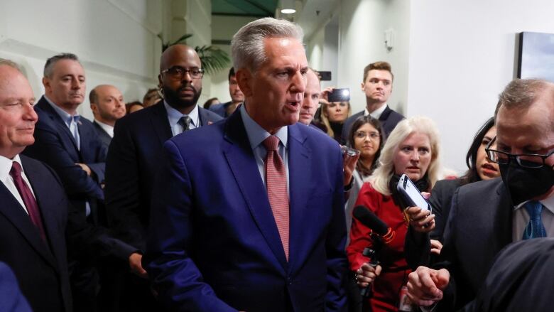 Grey-haired man stands at the centre of a group of reporters looking frustrated as he speaks.