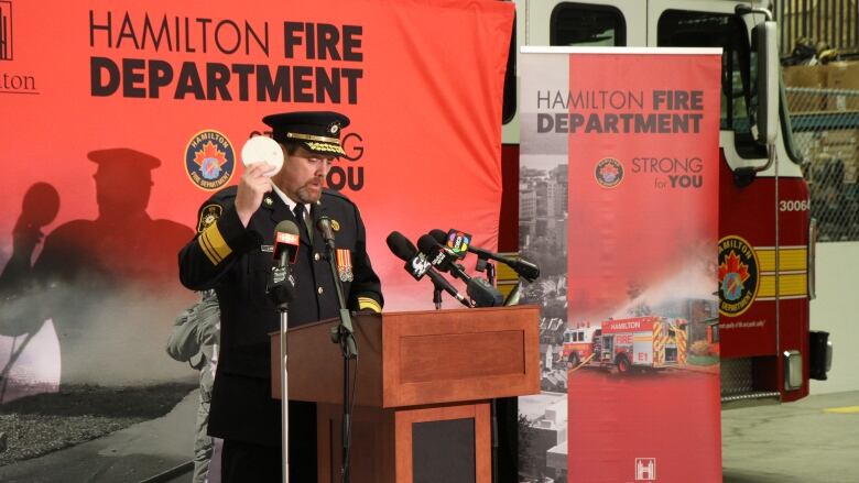 A firefighter in a suit holds up a fire detector. He is in front of a fire truck. 