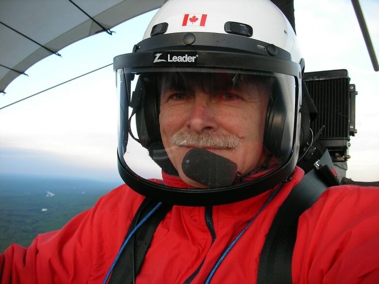 A man with moustache and a pilot helmet in red jacket, with an aircraft wing and a hill in the background.