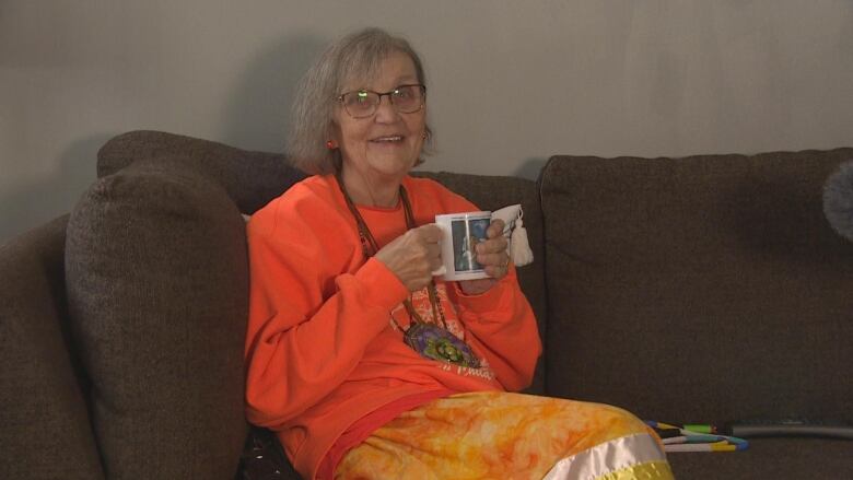 Knowledge keeper Caroyln Moar sits on a couch while wearing an orange sweater and orange, white and yellow ribbon skirt.