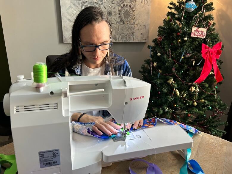 A person with long hair and glasses sews colour fabric with a Singer sewing machine.