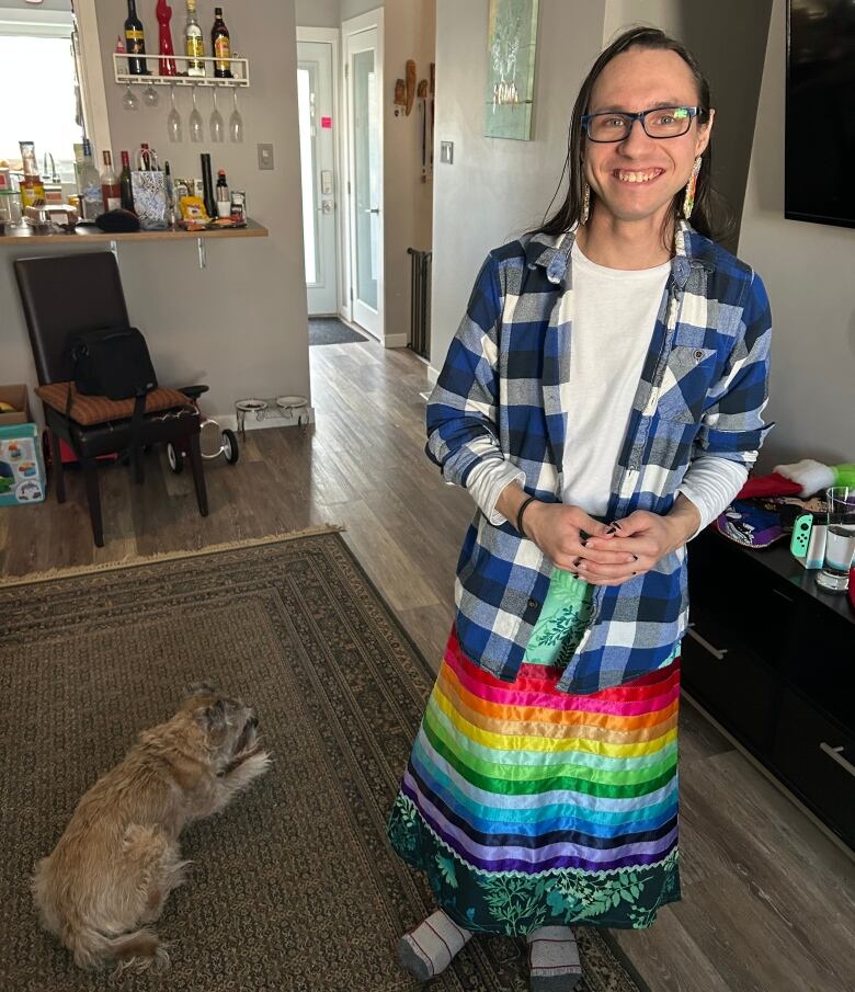 A two-spirit person with long hair and glasses stands in a room, wearing a long, colourful skirt with a rainbow of ribbons.