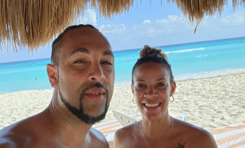 A man and woman stand under a shack holding drinks at a beach.