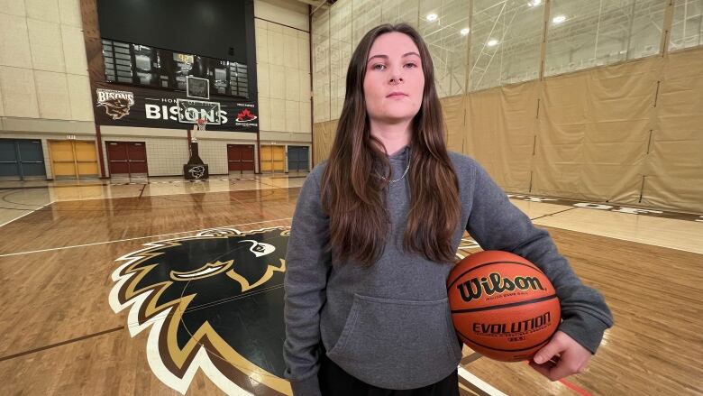 A woman stands in a university gymnasium with a basketball under her arm.