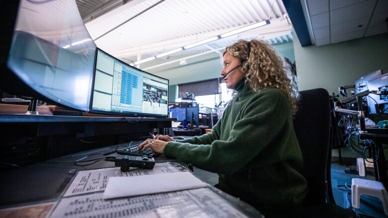 A woman sits in front of a computer with a headset and several monitors in front of her.