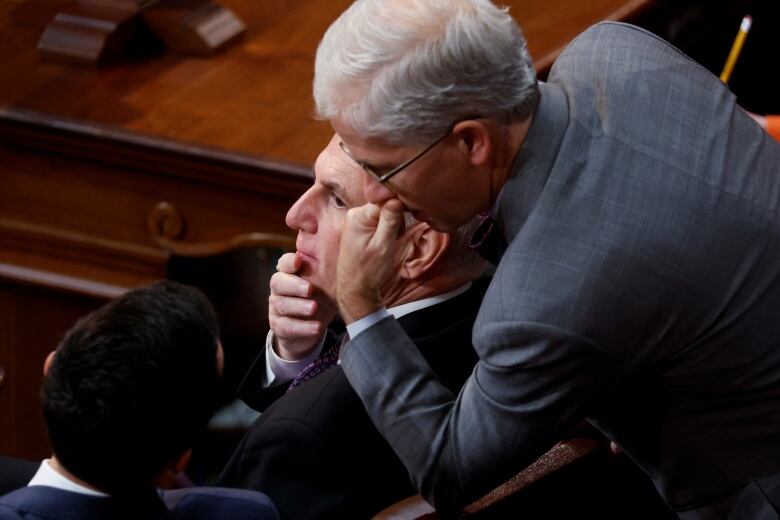 A grey-haired man leans over to communicate with another serious-looking grey haired man. 