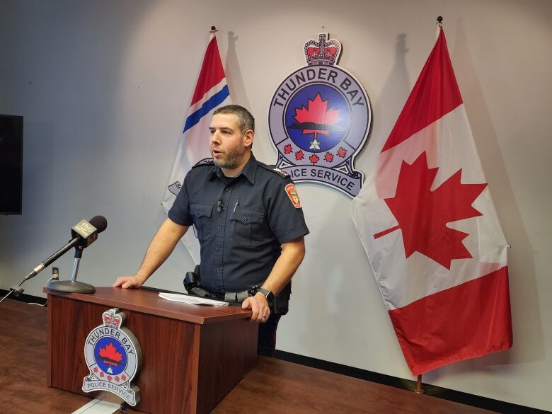 A police officer stands at a podium speaking into a microphone with the Canadian flag and Thunder Bay police logo behind him.