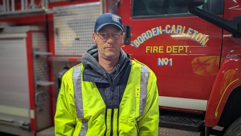 Shawn Jessome, the Borden Carleton fire chief stands in front of a red firetruck for a photo. He is wearing a blue cap, glasses and is not smiling. 