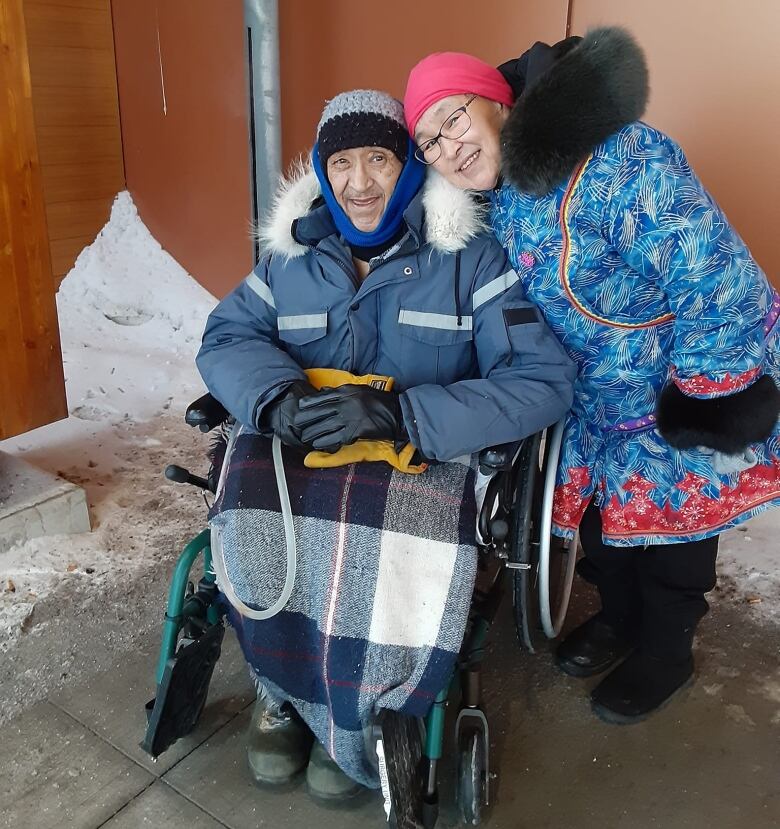 A man in a parka and blanket sits outside a building in a wheelchair with a smiling woman standing beside him with her arm around him.   