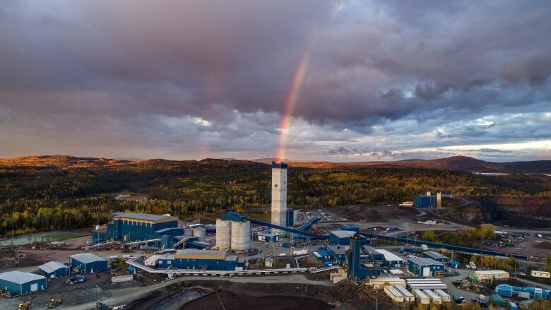 A rainbow arches down out of a cloudy sky to a gold mine set amongst the forested hills of northern Ontario. 