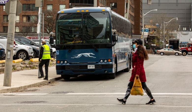 A Greyhound bus sits parked on a side street as a person in a mask walks past. An employee in a high-vis vest stands near the blue bus.