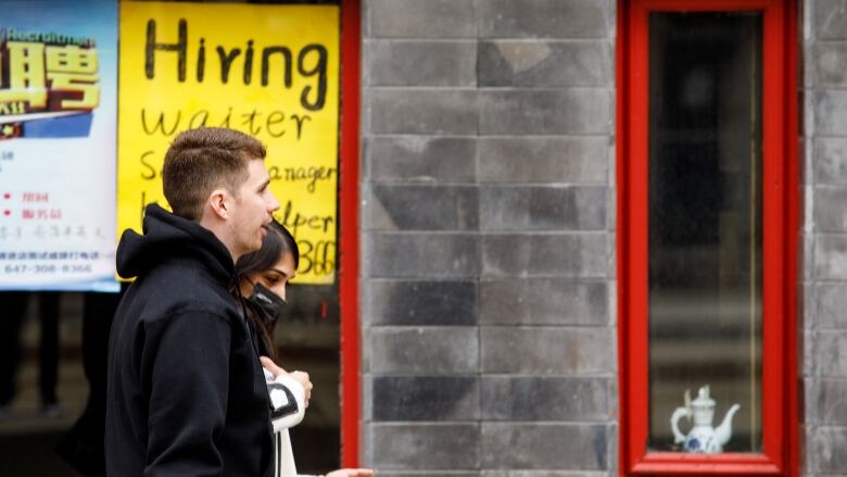 A man and a woman walk by a sign that says 'hiring waiter.'