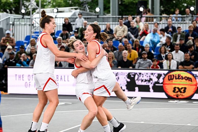 Three basketball players wearing white jerseys embrace in celebration at mid-court.