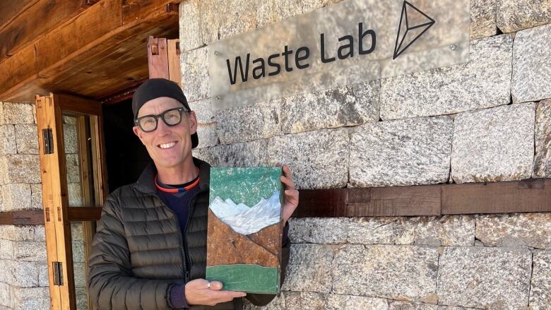 A man, smiling at the camera holding a painting-like piece of a mountain range made with scrap metal.