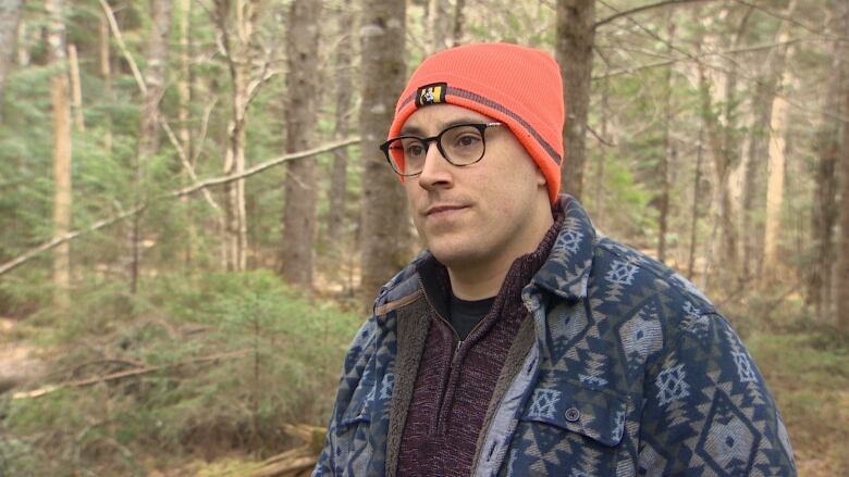 A man in a hunter's orange knit hat and glasses stands in the forest. He is also wearing a blue sweater.