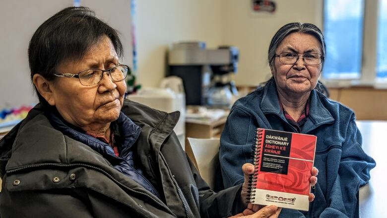 A woman is sitting down, holding a small booklet while a second woman is sitting next to her. 