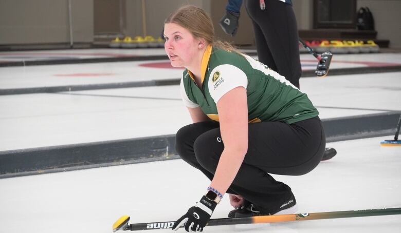 A woman squats on the pebbled ice with a broom in her left hand.