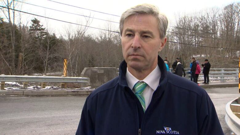 A man is seen standing near a bridge. A group of people are seen standing on the sidewalk of the bridge in the background.