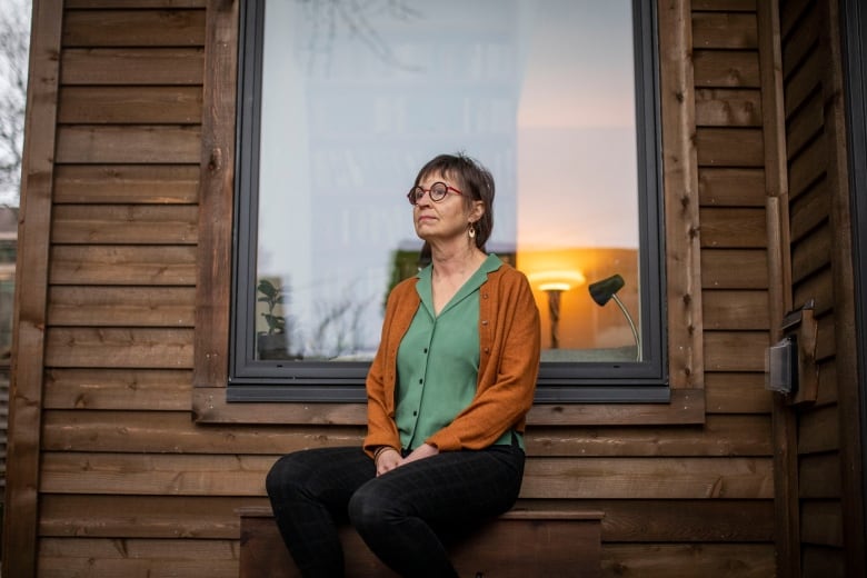 A woman sits outside the wood-framed window of a laneway home, looking away from the camera.