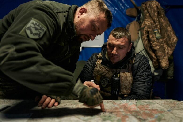 Two men in military uniforms. One sits, the other stands with his finger on the map, which is laid out flat.