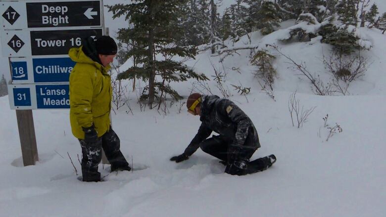 Posed in front of ski-slope signage, a man wearing yellow and another wearing black look at the depth of the snow under their feet.