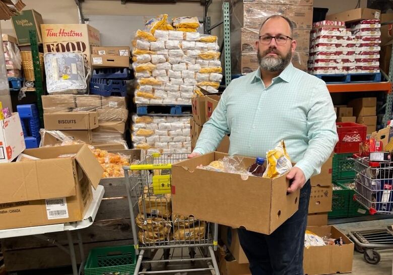 A bearded man with glasses holds a large cardboard box in a storage room. Behind him are shelves stocked with food products. 