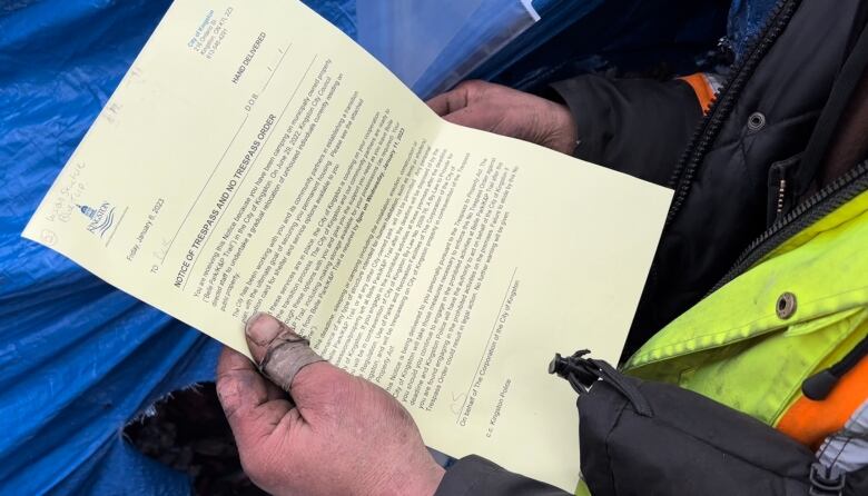 A man with hands covered in dirt and tape holds a yellow piece of paper with the logo for the City of Kingston on it.