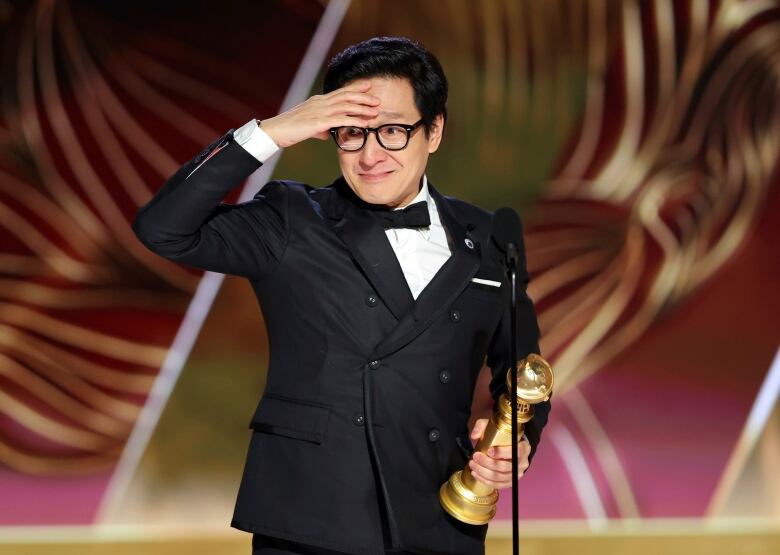 A man in a tuxedo holds a golden statuette in one hand, bringing his other to his forehead, while standing in front of a microphone.