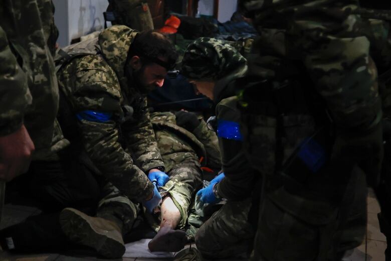 The two servicepeople are in military fatigues, wearing blue gloves and blue armbands. One uses scissors to cut away the patient's pantleg. 