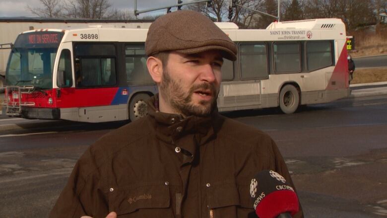 A man is seen from the shoulders up. He's wearing a brown coat and a newsboy cap. Behind him is a large bus. 