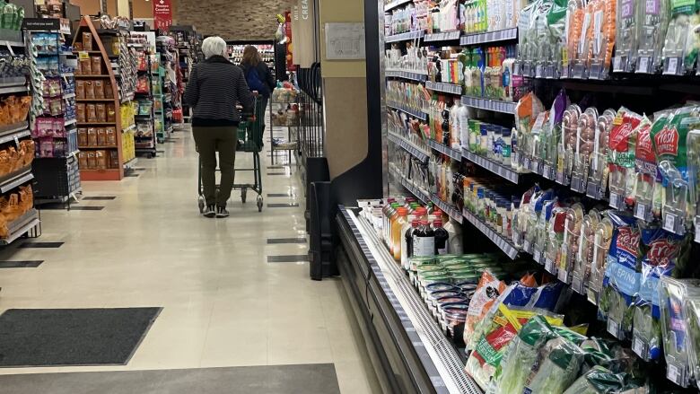 An aisle at a grocery store shows salads and other green items, with a person pushing a cart off in the distance.