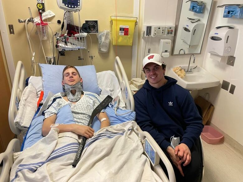 Two men sit in a hospital room. One is lying in a hospital bed, with a brace around his neck, and is holding a sledge hockey stick. The other is sitting on the bedside. Both are smiling toward the camera.