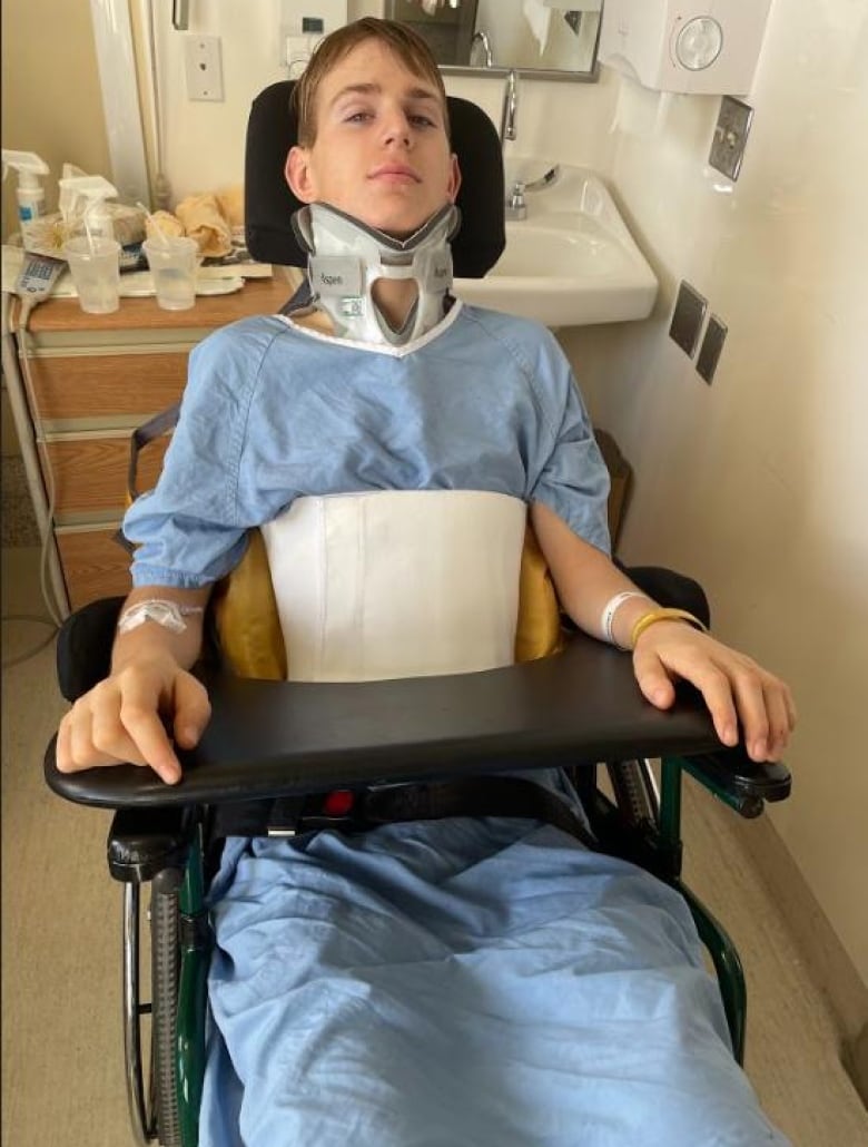 A boy, wearing a hospital gown and a beck brace, sits in a wheel chair in a hospital room.