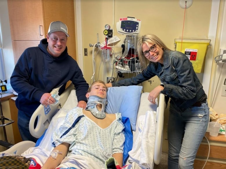 A boy is lying in a hospital bed, with a neck brace, with his parents standing next to the bed.