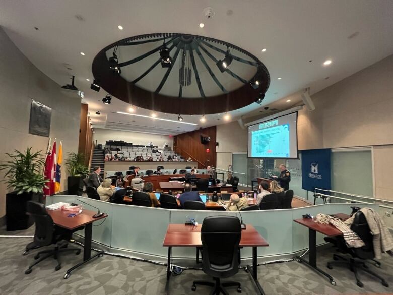 People sitting in an auditorium and around a horse-shoe shaped table watching a man speak at a podium.