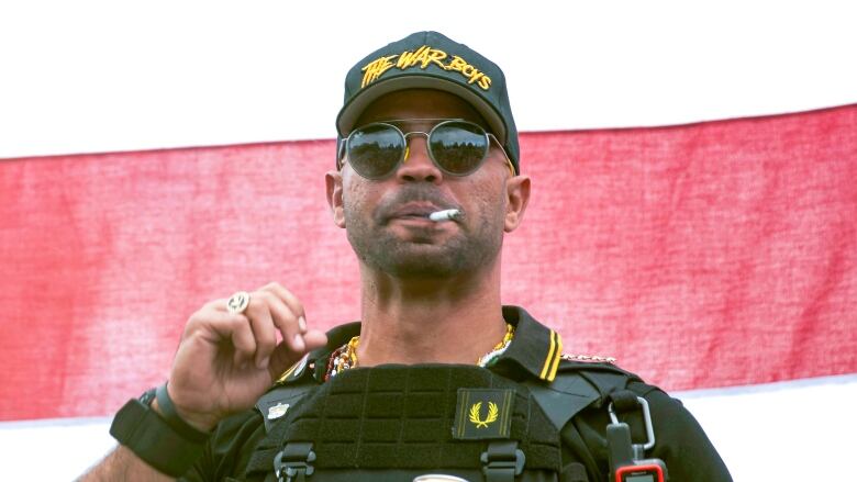 A man in a baseball cap and military gear smokes a cigarette while his vest holds a White Claw beverage.