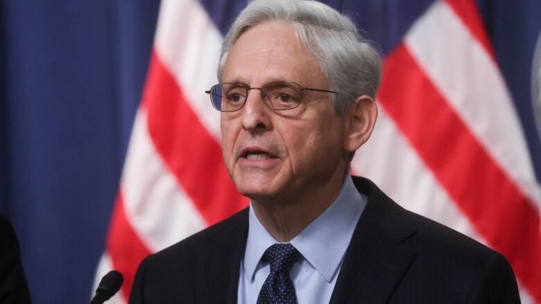 Grey haired man with glasses speaking, with a U.S. flag behind him. 