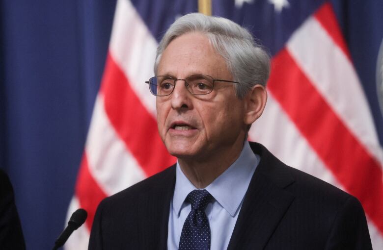 Grey haired man with glasses speaking, with a U.S. flag behind him. 