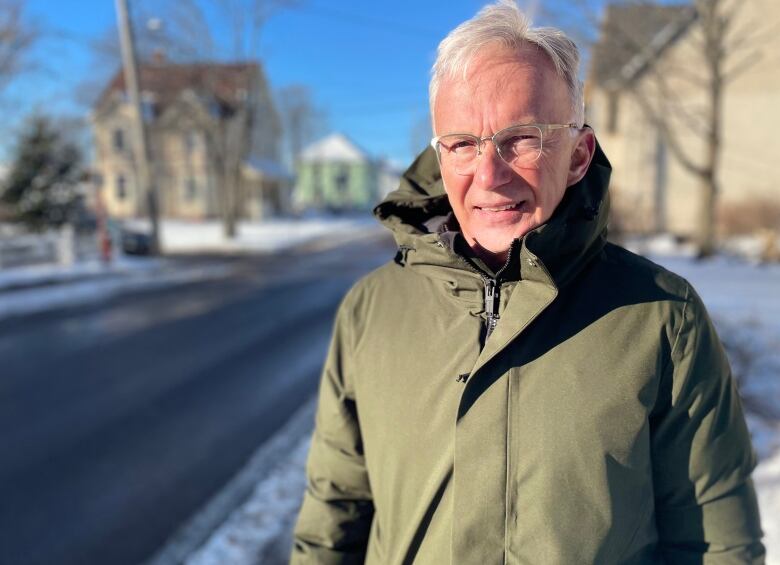 A man wearing a green parka stands on a snow-covered sidewalk. 