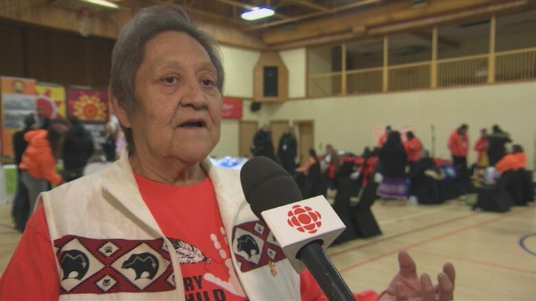 An old Indigenous woman, with short grey hear, is wearing a white vest is a red strip across the chest. Inside the red stride is a diamond pattern with bears illustrated in an Indigenous style. She's wearing an orange 'Every Child Matters' shirt. The woman is talking into a microphone.