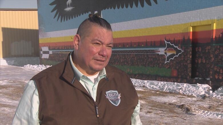 An Indigenous man, wearing a brown vest with a patch sewn on the left chest and a mint-green dress shirt underneath, is standing outside in front of a colourful brick. The wall has a large arrow painted on it, as well as earth, trees and birds.