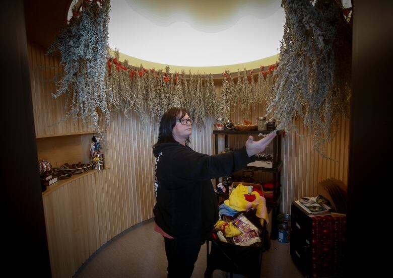 A woman stands in a circular room with drying plants hanging from the ceiling, one hand raised to touch a bunch of sage.